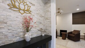 Un muro de piedra con un diseño de loto, una consola negra, un florero con flores rosadas, una pequeña escultura y una zona de estar con una silla marrón y una mesa redonda.