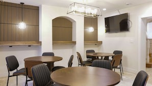 Un salón de conferencias con mesas redondas, sillas, un televisor montado y una lámpara en el techo.