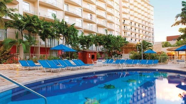A hotel pool area with blue lounge chairs, a clear pool, and a multi-story building with balconies. - Spacious 2-BR/2-bath suite with full kitchen & living room, minutes from beach (Honolulu)