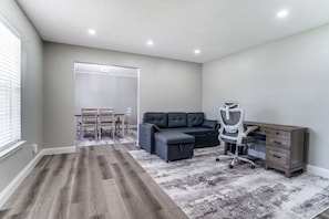 Ein Zimmer mit grauem Sofa, einem Schreibtisch mit Schubladen, einem Stuhl und einem gemusterten Teppich.