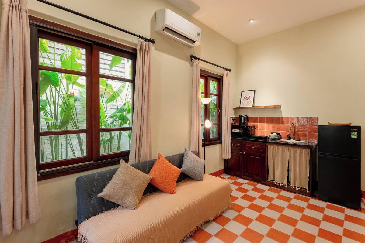 A room with a sofa, a kitchen area, and large windows with curtains.