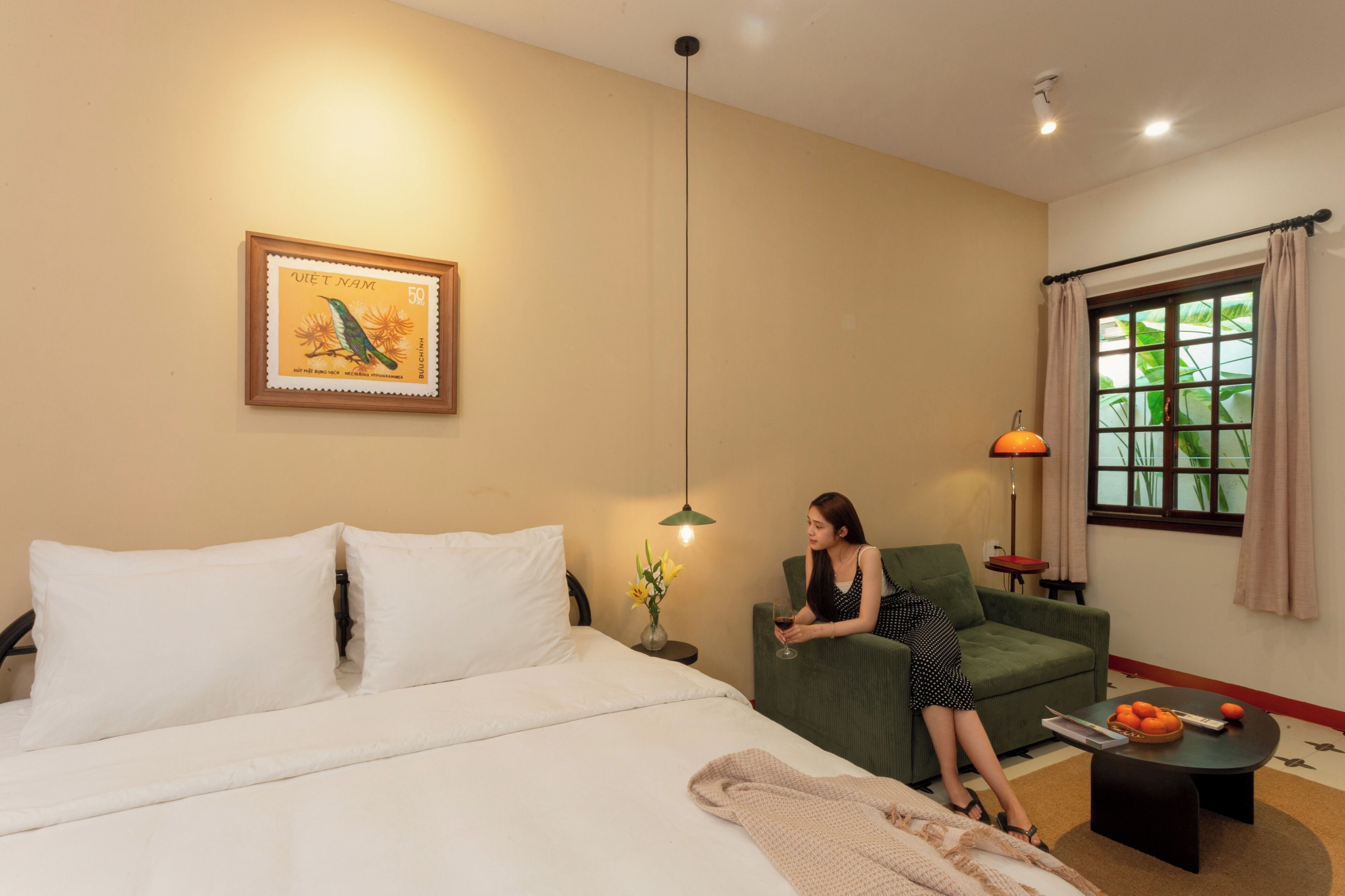 Una habitación de hotel con cama, un sillón verde, una mesita con lámpara y una ventana con cortinas.