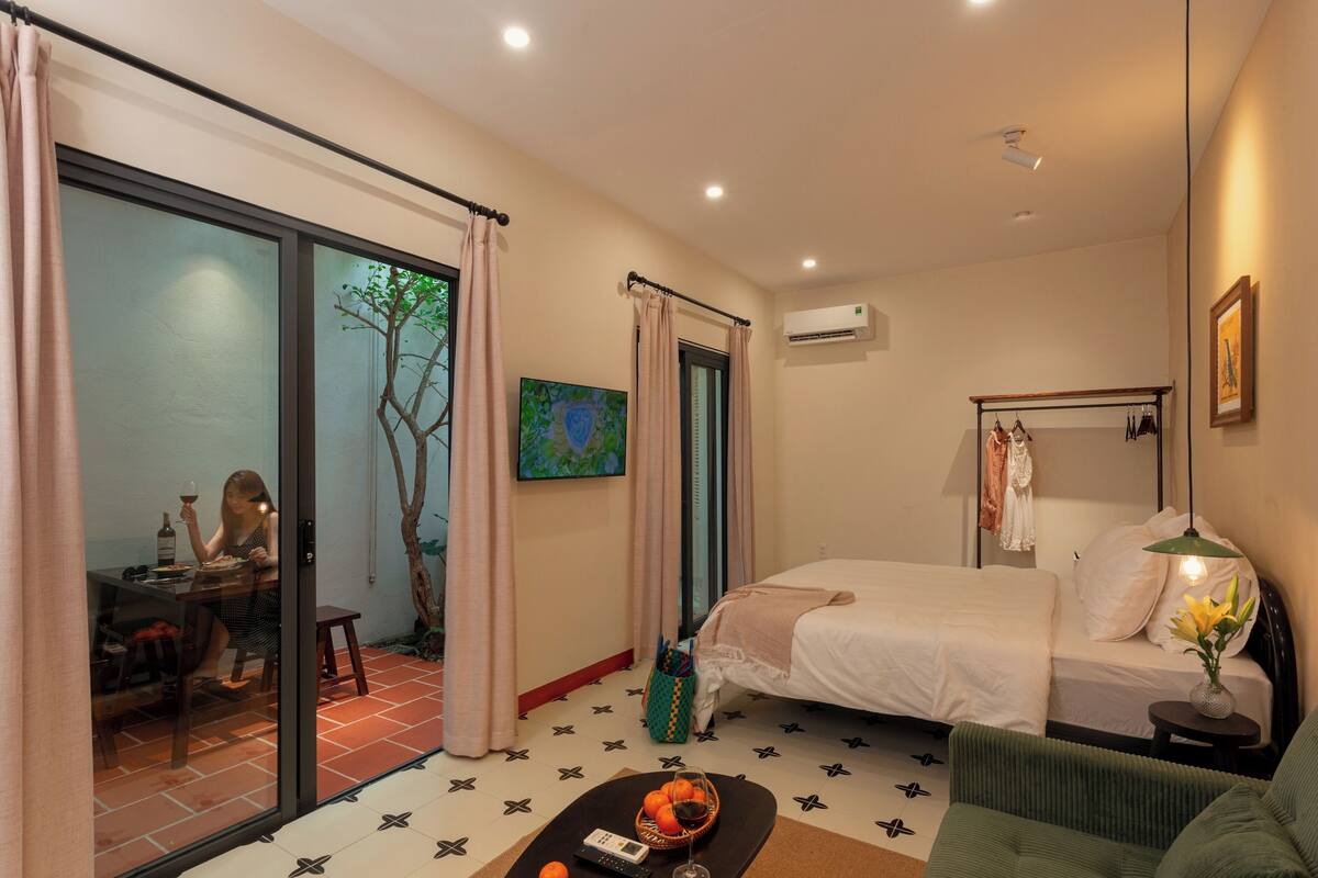 A bedroom with a bed, a sofa, a dining table, and a sliding glass door leading to a balcony.