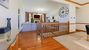 A modern living room with a wooden staircase, a large wall clock, and a flat-screen TV displaying 'NETFLIX'.