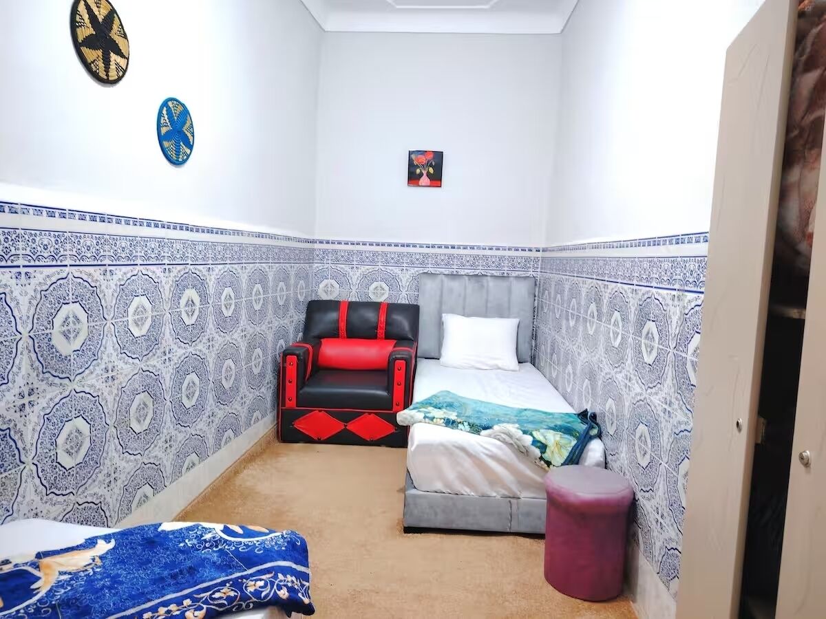 A room with a bed, a chair, a small stool, and a wall with patterned tiles.