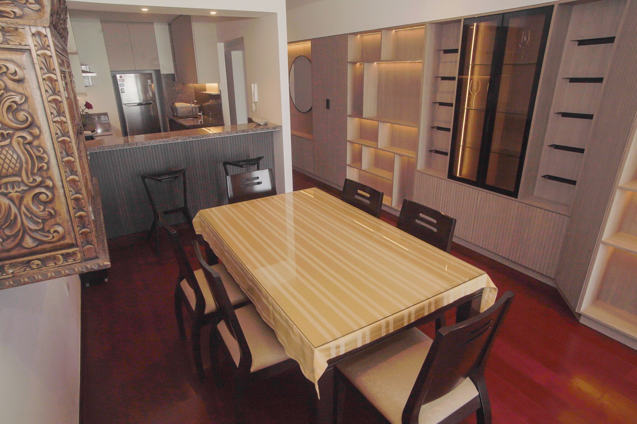 Un comedor con mantel a rayas, sillas de madera, una cocina al fondo y una pared decorativa con estanterías.