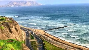 Una carretera costera con vista al acantilado, un camino serpenteando a lo largo de la costa y una cadena montañosa distante.