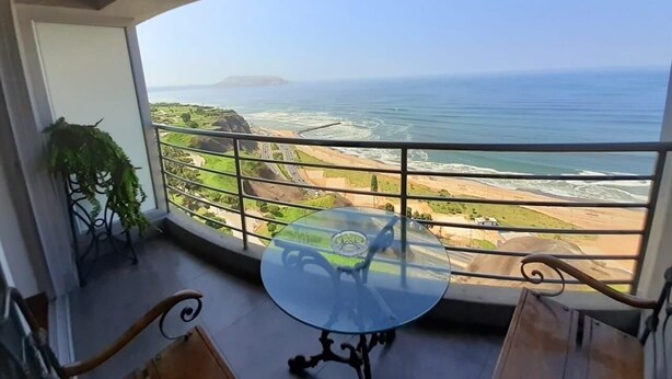 Un balcón con una mesa y sillas de vidrio, con vistas a un paisaje costero y al mar, con tierra a la distancia.