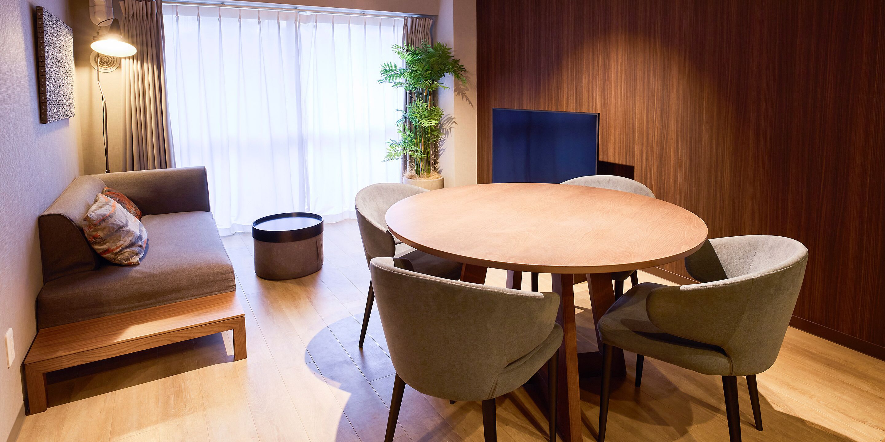A modern room with a round wooden dining table, four chairs, a sofa with a cushion, a floor lamp, and a potted plant.