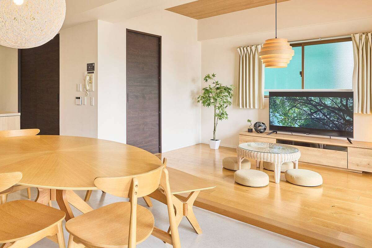 A modern kitchen with a round dining table, wooden chairs, a built-in oven, and a large window with curtains.