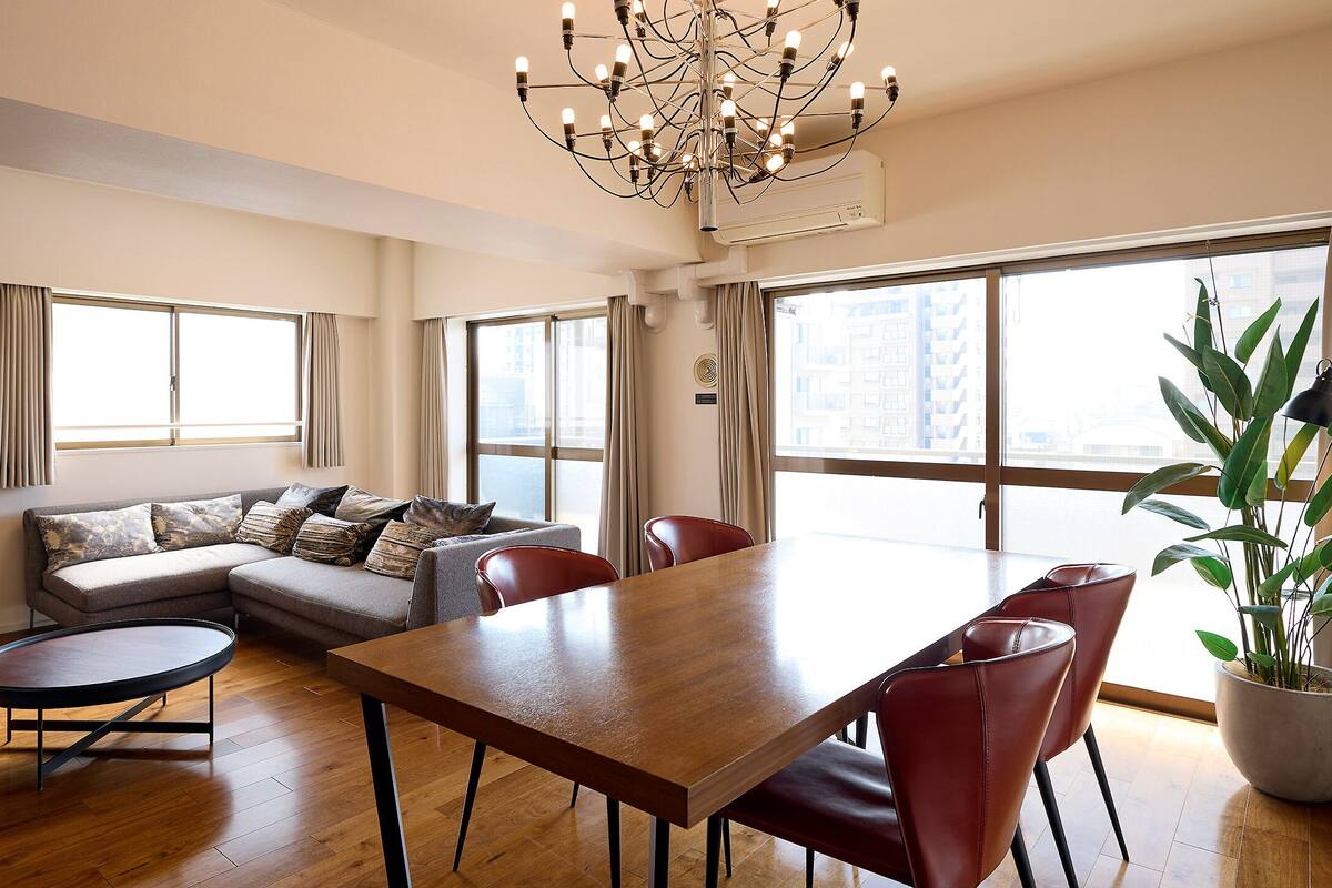 A modern living room with a sofa, dining table, chairs, a potted plant, and large windows.