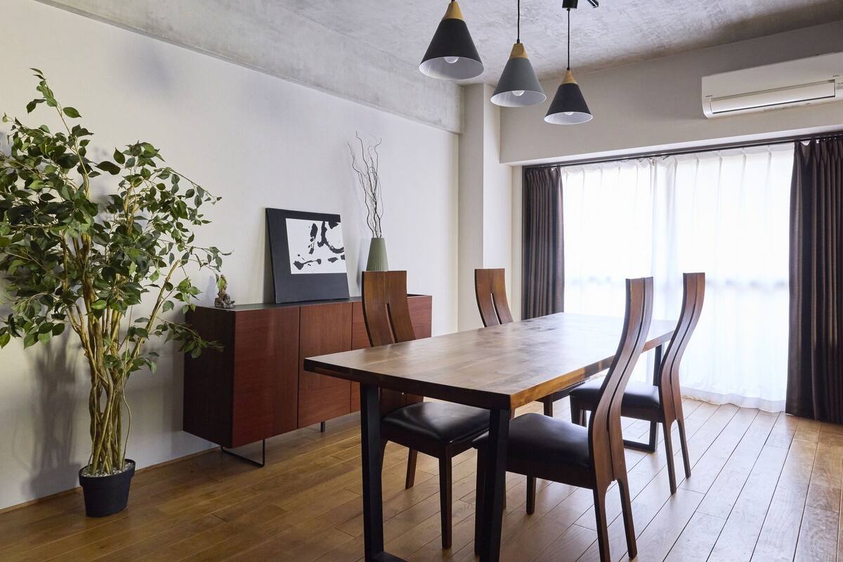 A dining area with a wooden table, chairs, a potted plant, a cabinet, and a large window with curtains.