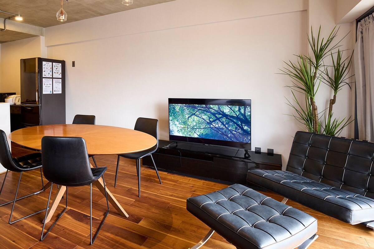 A modern living room with a wooden floor, a round dining table, black chairs, a black sofa, a television, and a potted plant.