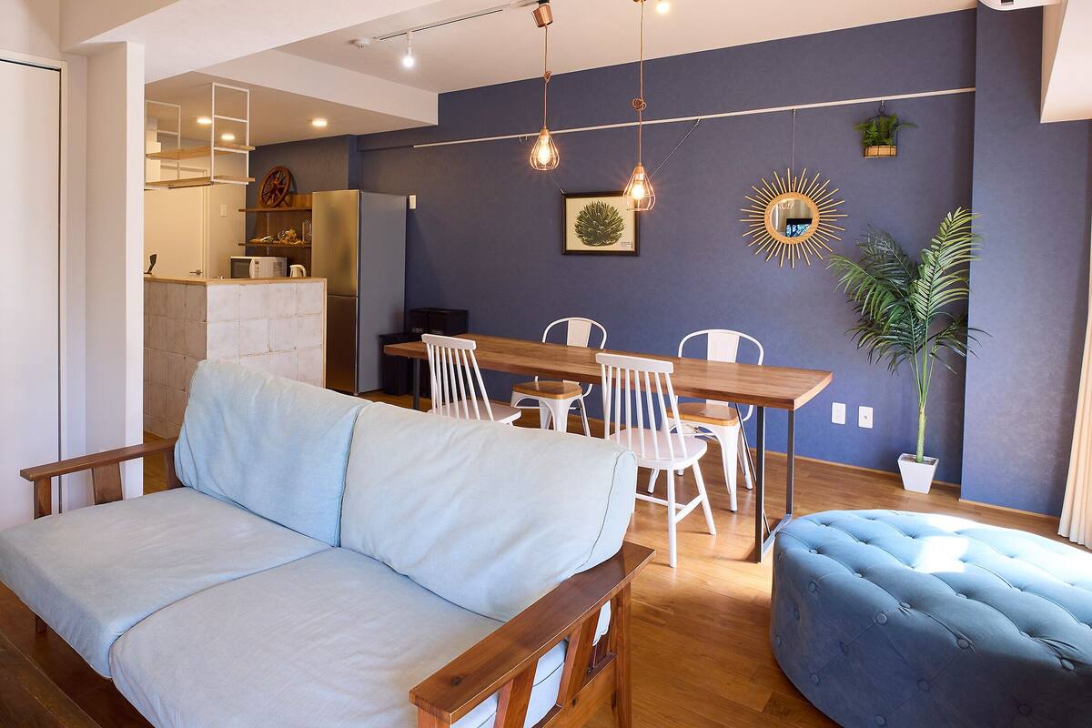 A modern living room with a sofa, ottoman, dining table, chairs, and a kitchen area in the background.