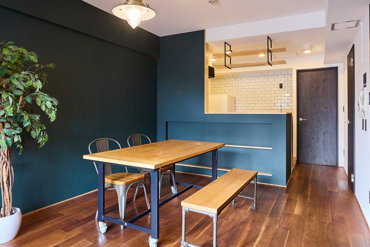 A dining area with a wooden table and chairs, a potted plant, and a kitchen in the background.