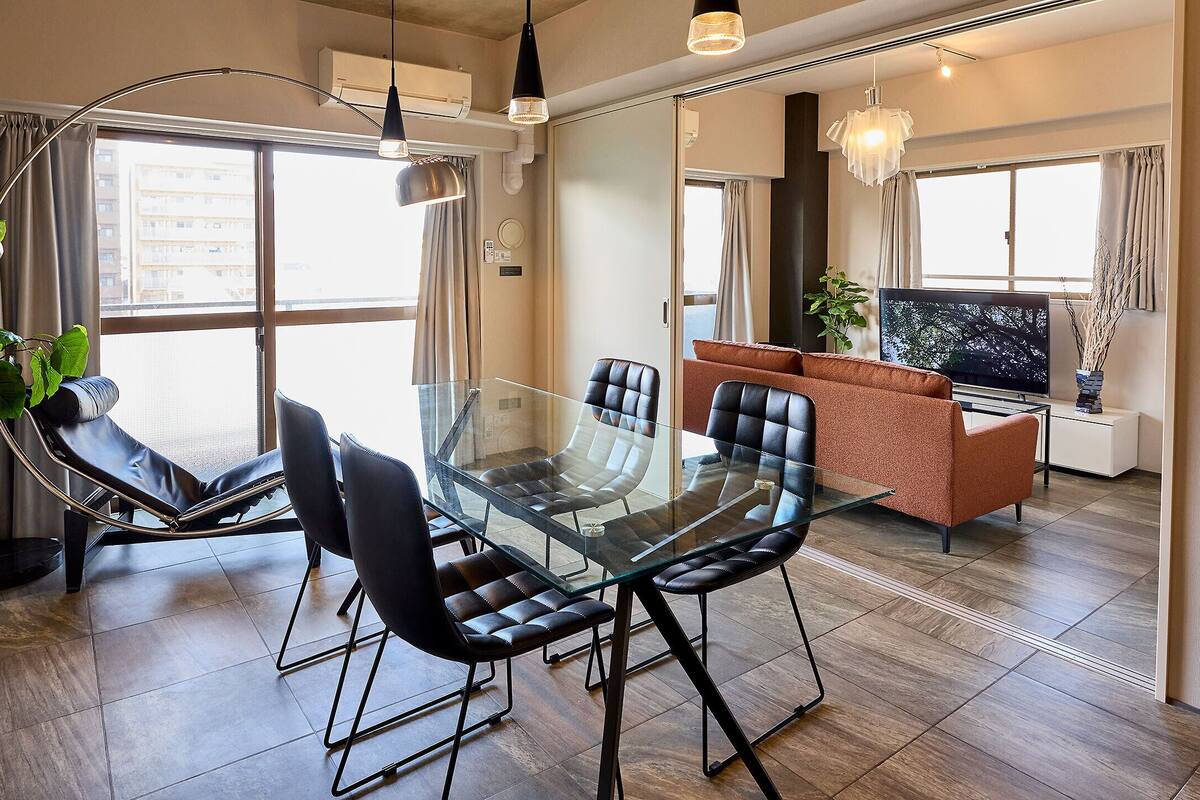 A modern dining area with a glass table and black chairs, a sofa, and a large window with curtains.