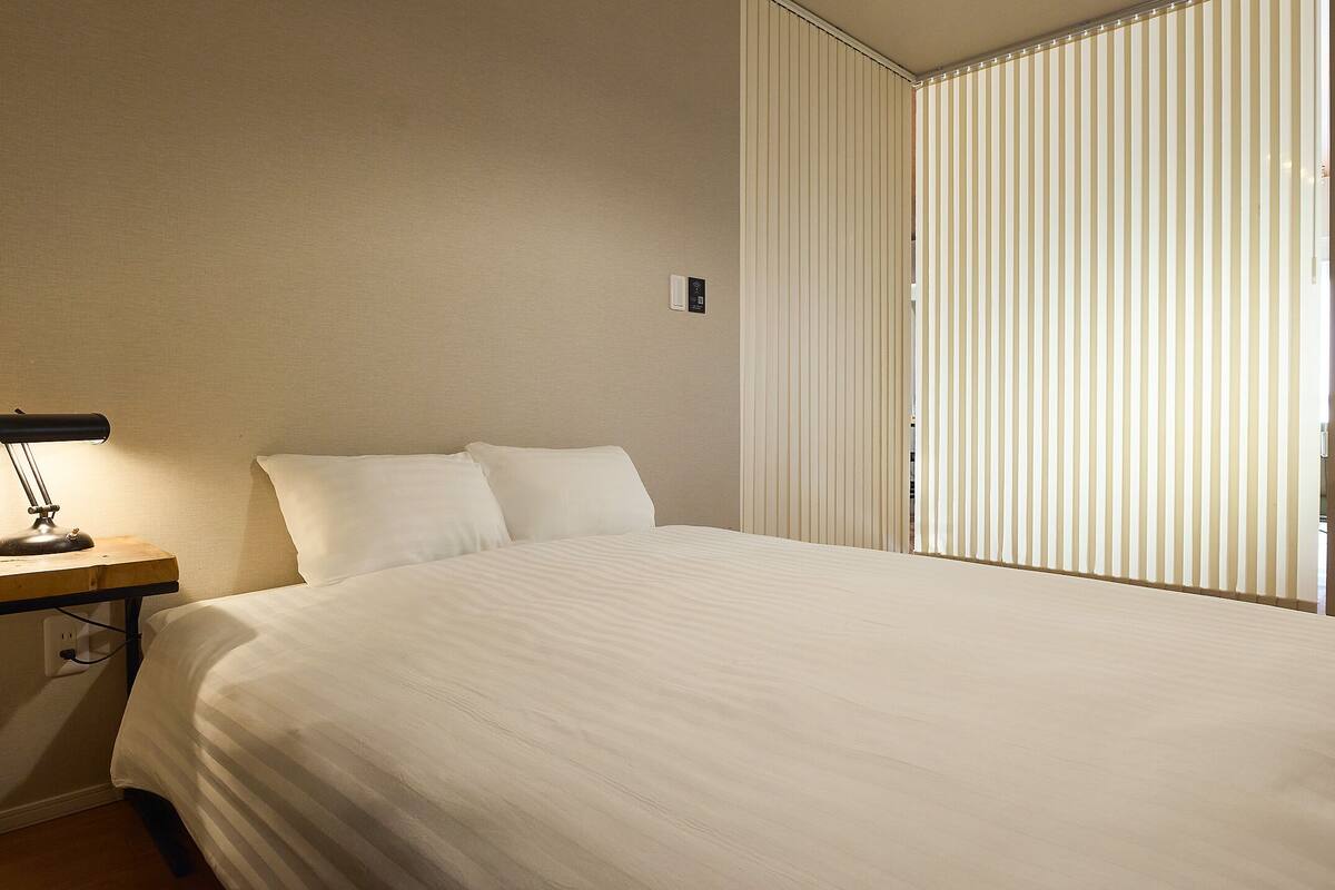 A neatly made bed with white bedding, a bedside table with a lamp, and a chair with a desk.