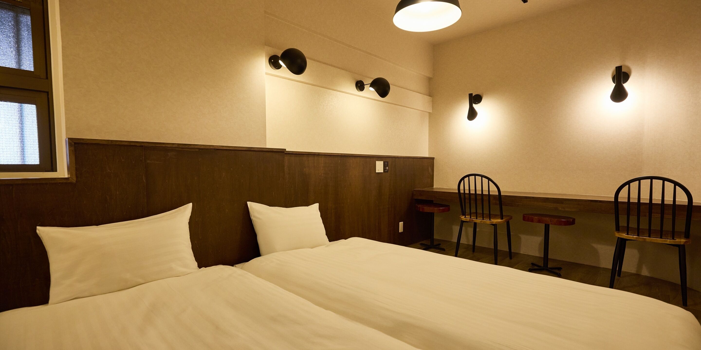 A hotel room with a large bed, two chairs, a small table, and wall-mounted light fixtures.