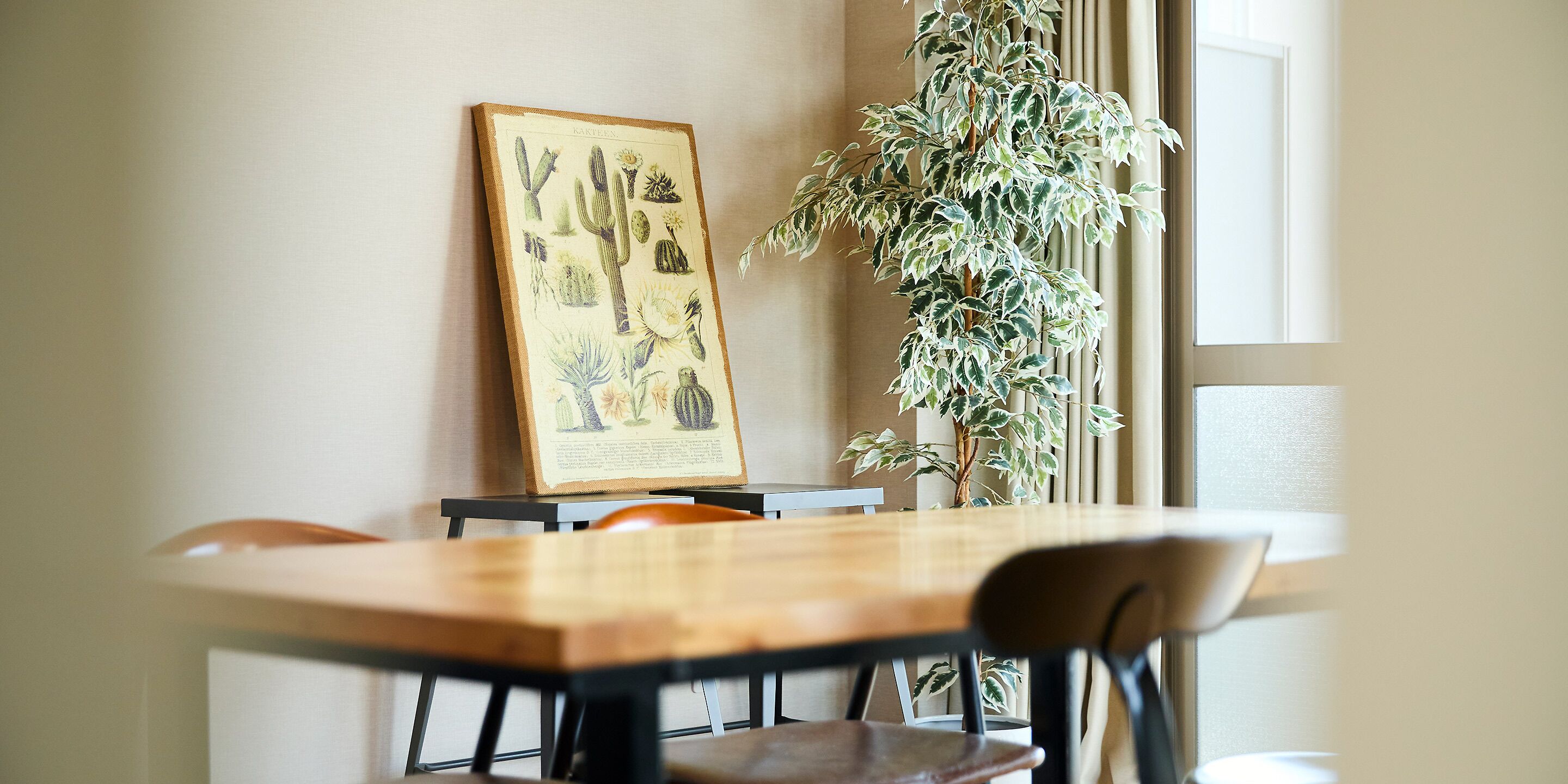 A dining area with a wooden table, chairs, a framed botanical illustration on the wall, and a potted plant.