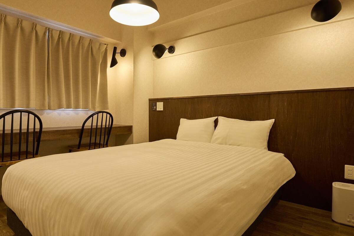A bedroom with a bed, two chairs, a window with curtains, a wall-mounted lamp, and a bedside table with a lamp.
