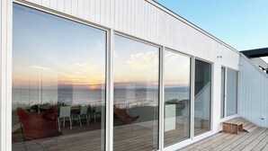A modern building with large glass windows, a wooden deck, and a view of the sea.