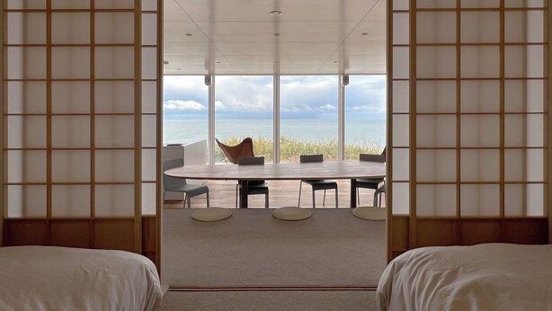 A room with a view of the sea, a round table with chairs, and two beds.