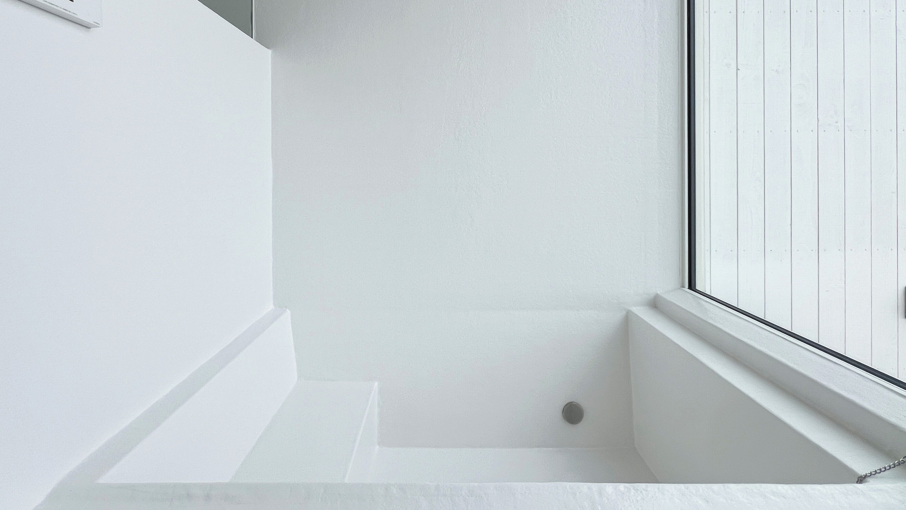 A white bathtub with a window, a white wall, and a small circular object on the floor.