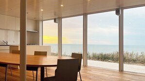A modern dining area with a large table and chairs, a kitchen in the background, and a view of the sea through large windows.