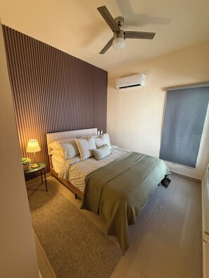 Un dormitorio con cama, ventilador de techo, aire acondicionado, ventana con persianas y una pequeña mesa con lámpara.