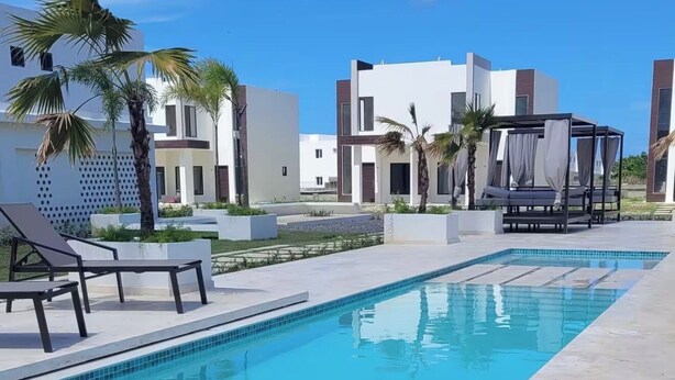 Une piscine avec des chaises longues, un espace de détente couvert et des bâtiments modernes en arrière-plan.