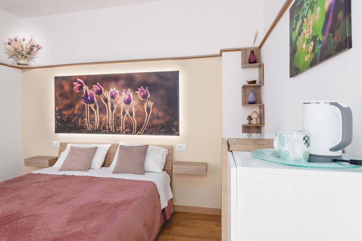 A bedroom with a bed, a nightstand, a coffee maker, and a wall-mounted shelf with decorative items.