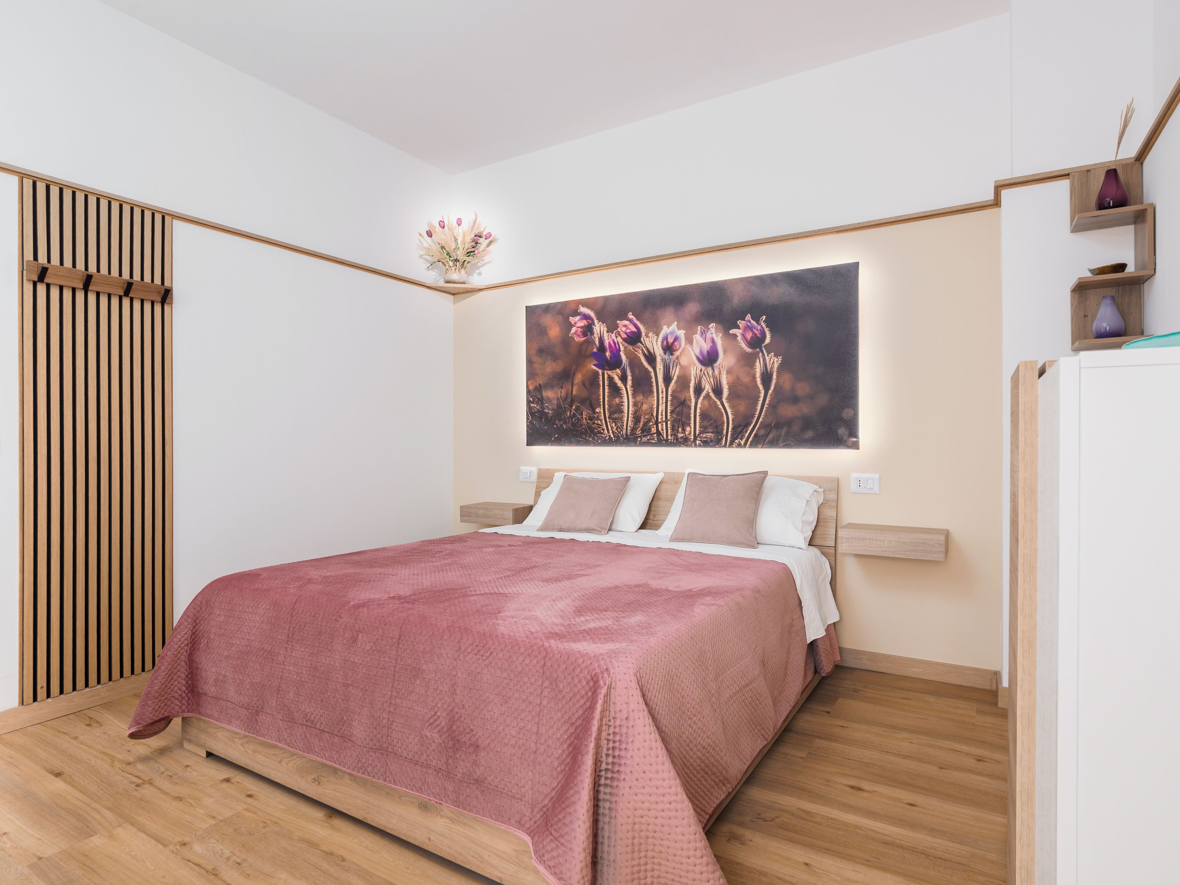 Een moderne slaapkamer met een houten bedframe, een roze sprei, een wandlamp en een groot bloemenkunstwerk boven het bed.