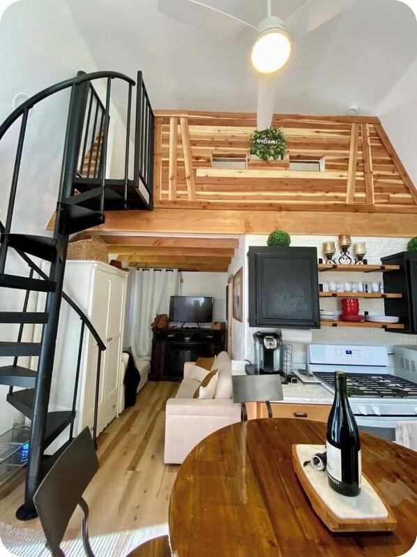 A loft-style living space with a wooden staircase, a dining area with a table and chairs, a kitchen with a stove, and a television in the background.