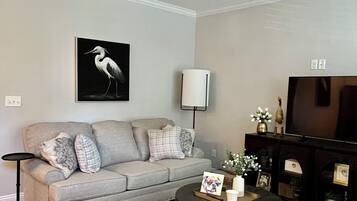 Uma sala de estar com ventilador de teto, um sofá cinza com almofadas decorativas, uma mesa de centro com porta-retrato e velas, um aparador com objetos decorativos e um tapete estampado.