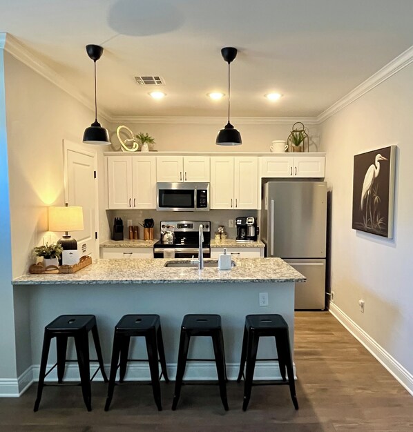 A modern kitchen with white cabinets, a stainless steel refrigerator, a microwave, and a coffee maker. There are three black bar stools in front of the counter.