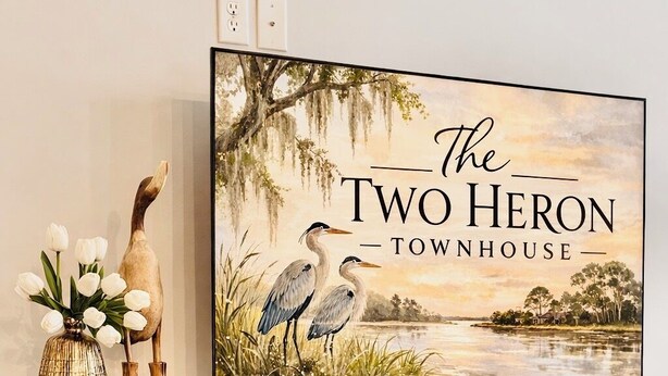 Ein Fernseher auf einem dunklen Holzschrank mit Glastüren, der eine Landschaft mit Vögeln zeigt und den Text 'The Two Heron Townhouse' anzeigt.