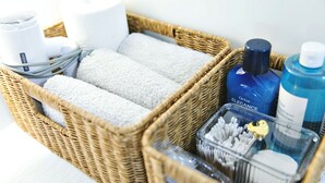 A wicker basket containing towels, a hairdryer, a bottle of Oriox Elegance, a blue bottle, and a container with cotton swabs.