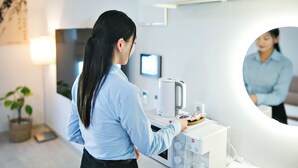 A woman in a blue shirt standing in a modern room with a coffee machine, a mirror, and a computer monitor on a desk.