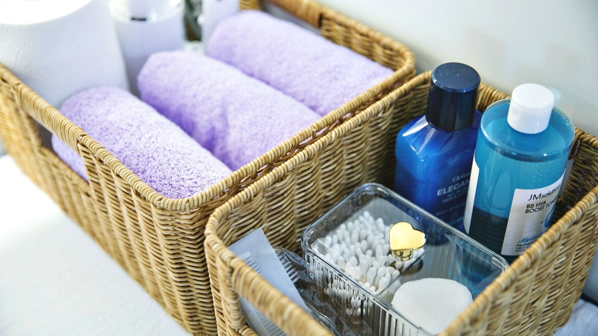 A wicker basket with lavender towels, a blue bottle, a clear container with white beads, and a gold-colored object.