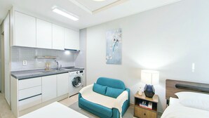 A compact hotel room with a kitchenette, a washing machine, a teal sofa, a bedside table with a lamp, and a bed.