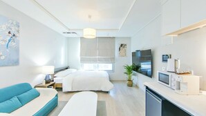 A modern hotel room with a bed, a sofa, a TV, a coffee machine, and a plant.