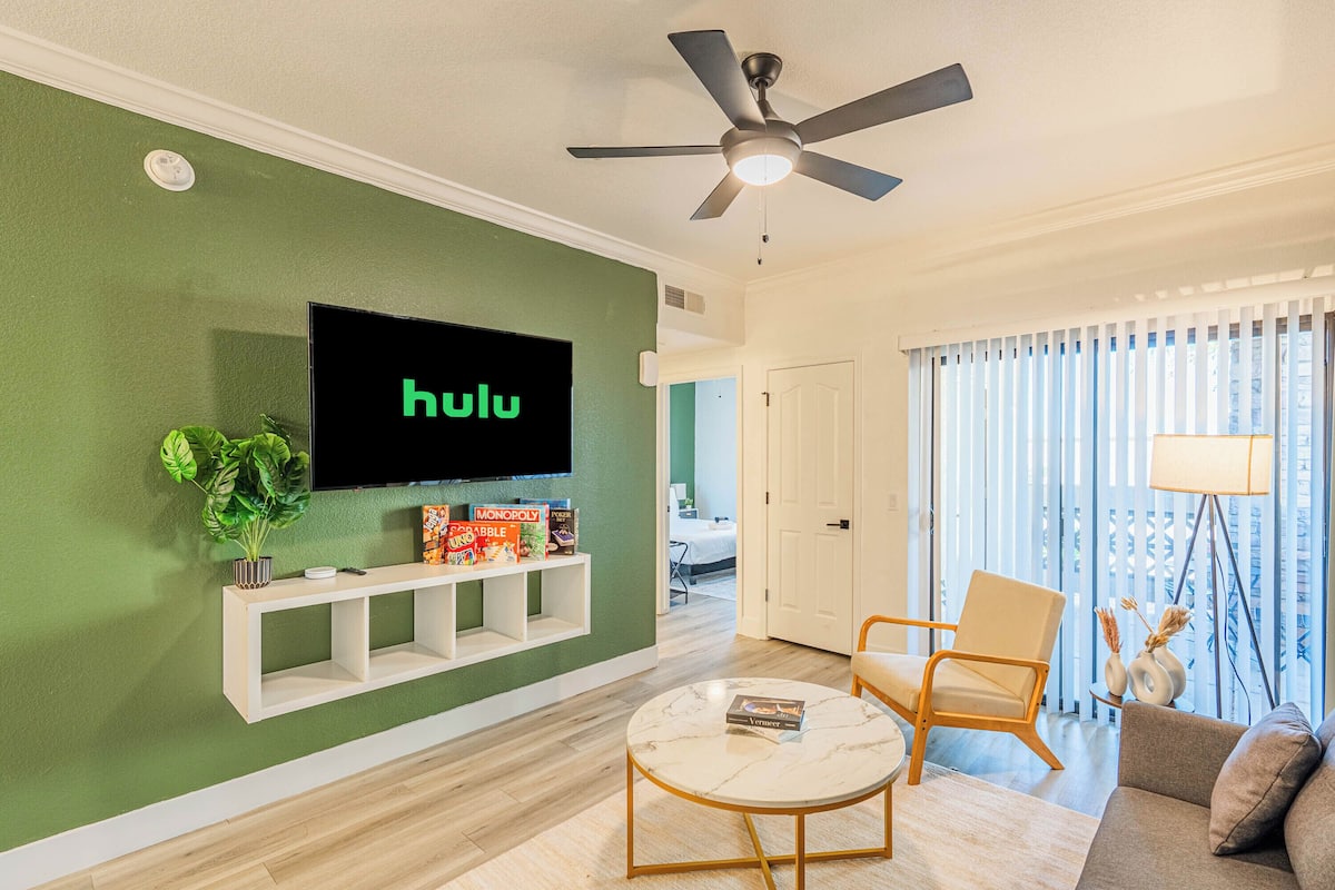 Un salon comprenant un téléviseur fixé au mur, un ventilateur de plafond, un canapé, une table basse et une plante.