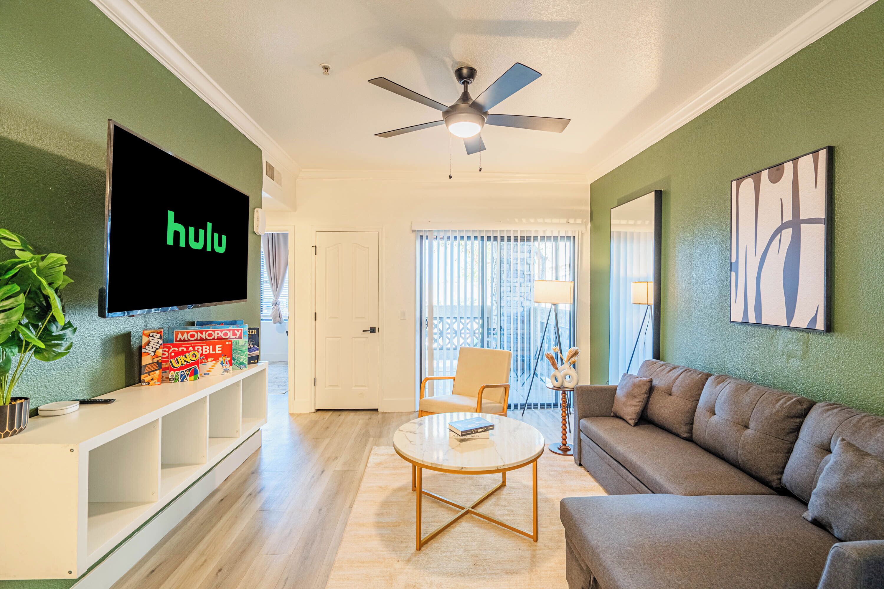 Una sala de estar con televisor de pantalla plana, ventilador de techo, sofá, mesa de centro y estantería con juegos de mesa.