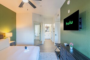 A hotel room with a bed, a TV mounted on the wall, a desk with a lamp, and a hallway leading to a bathroom. - San Portella (Tempe)