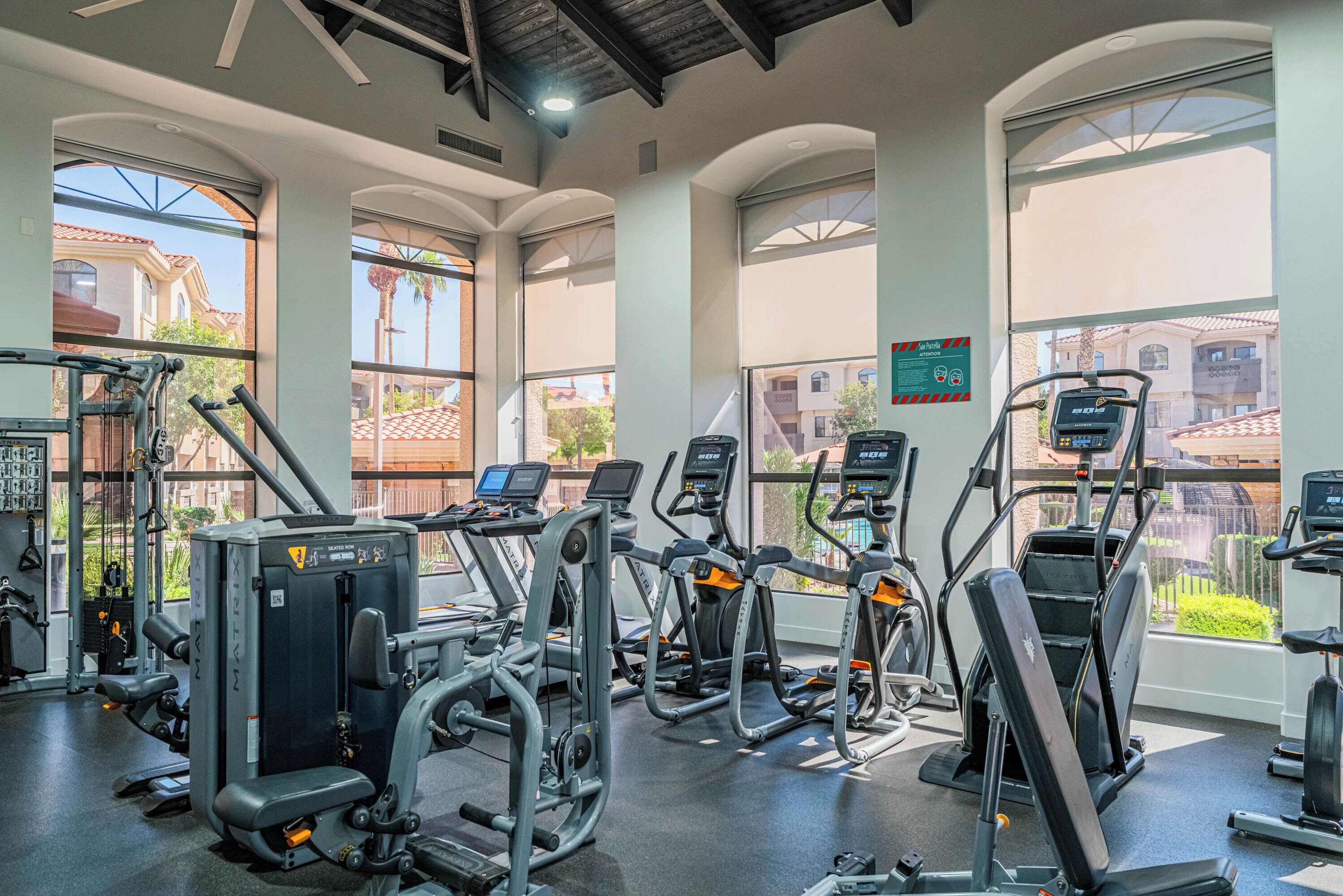 A gym with various exercise machines, large windows, and a view of buildings outside.