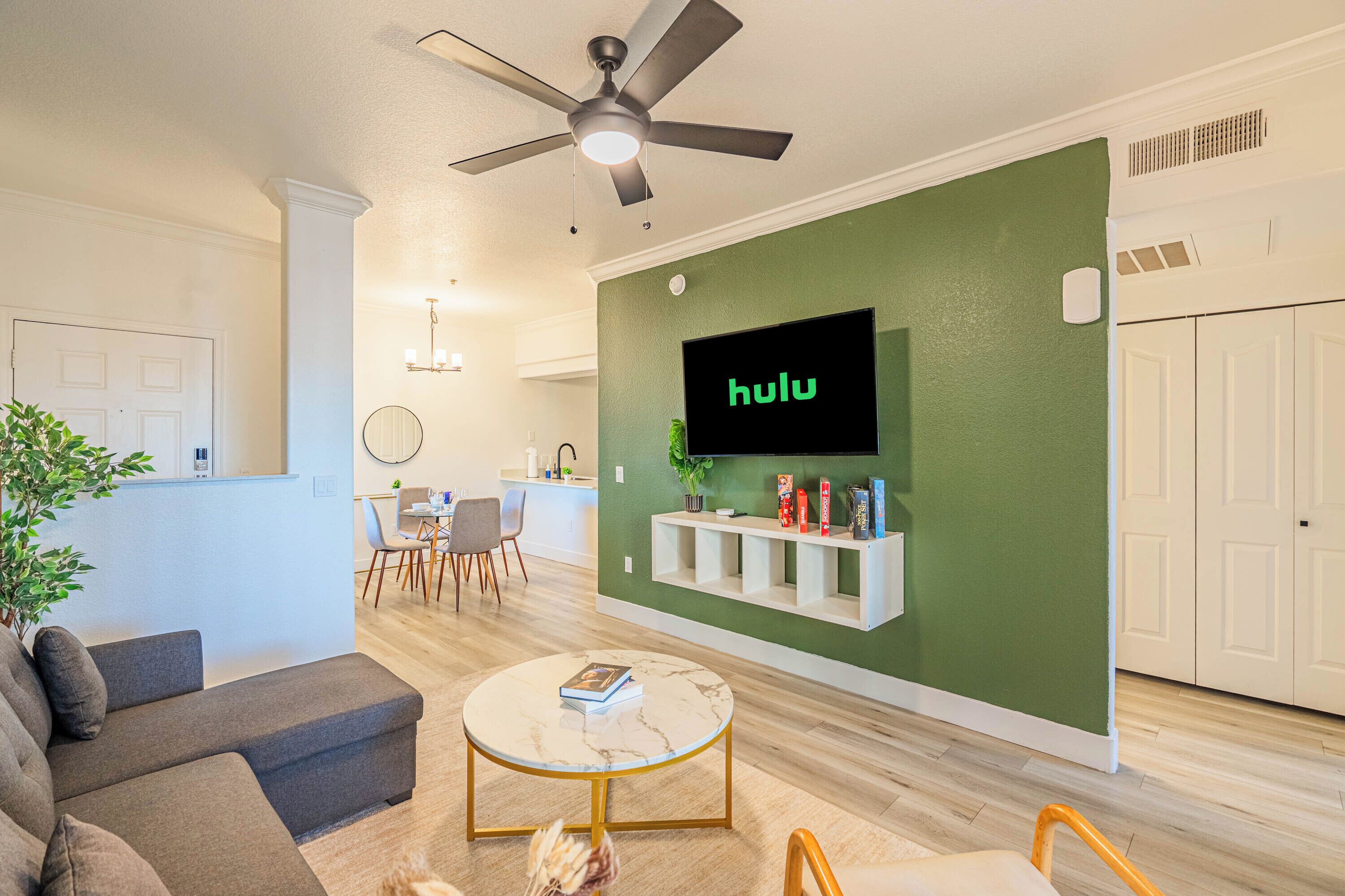 A living room with a sofa, a coffee table, a TV mounted on a green wall, a ceiling fan, and a dining area in the background.
