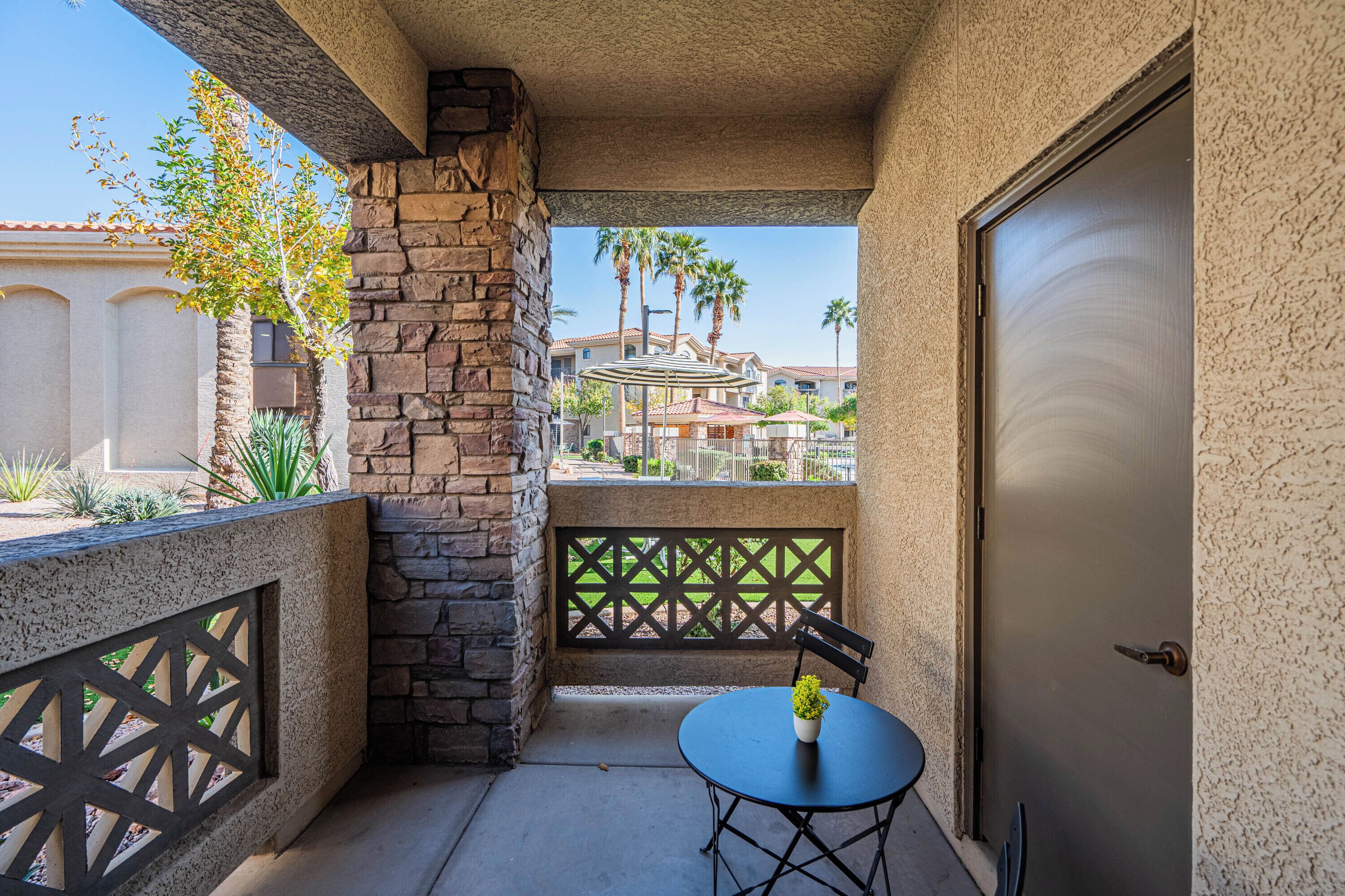 A balcony with a stone pillar, a table with a potted plant, and a view of palm trees and buildings in the background.