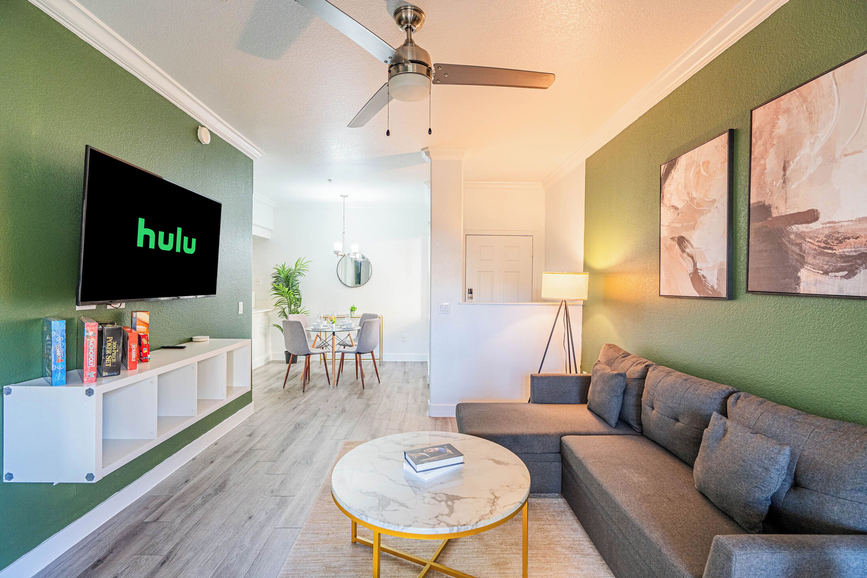 A living room with a flat-screen TV, a ceiling fan, a sofa, a coffee table, and a dining area with chairs and a table.