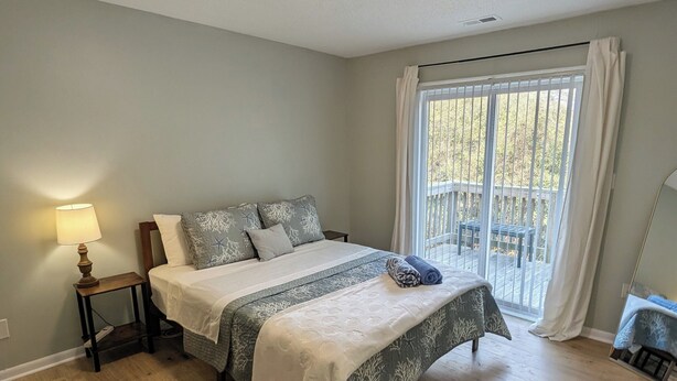 A bedroom with a bed, a nightstand, a lamp, a window with curtains, and a view of the outdoors. - Bright and cozy condo with inviting vibes for your home away from home (Morehead City)