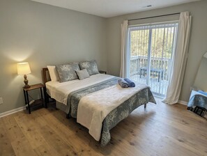 Et soveværelse med seng, natbord, lampe, et vindue med gardiner og udsigt over naturen.